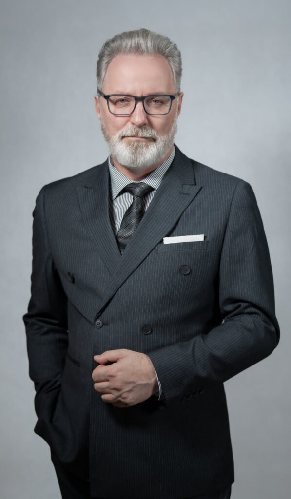 Mężczyzna w eleganckim garniturze, stojący. Nosi okulary, ma siwe włosy i zarost. Pozuje do zdjęcia na sesji biznesowej.