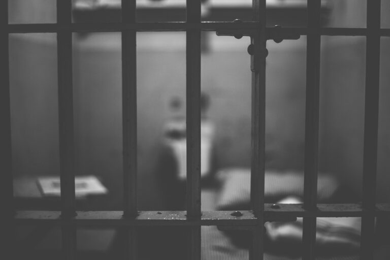 Czarno białe kraty celi więziennej, za nimi małe pomieszczenie dla więźnia.